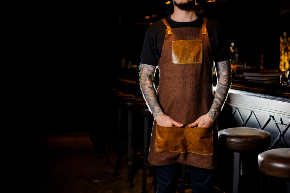 Tattooed Bartender Standing in front of Bar in Apron.